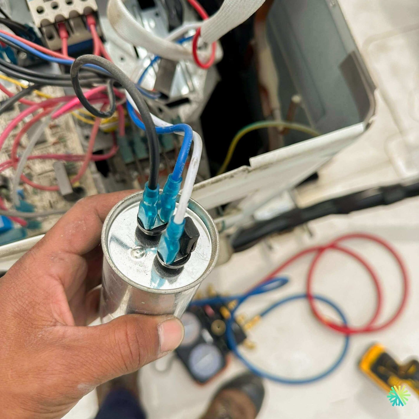 Hand holding electrical capacitor near wiring in HVAC repair setup with tools and wires visible in the background.