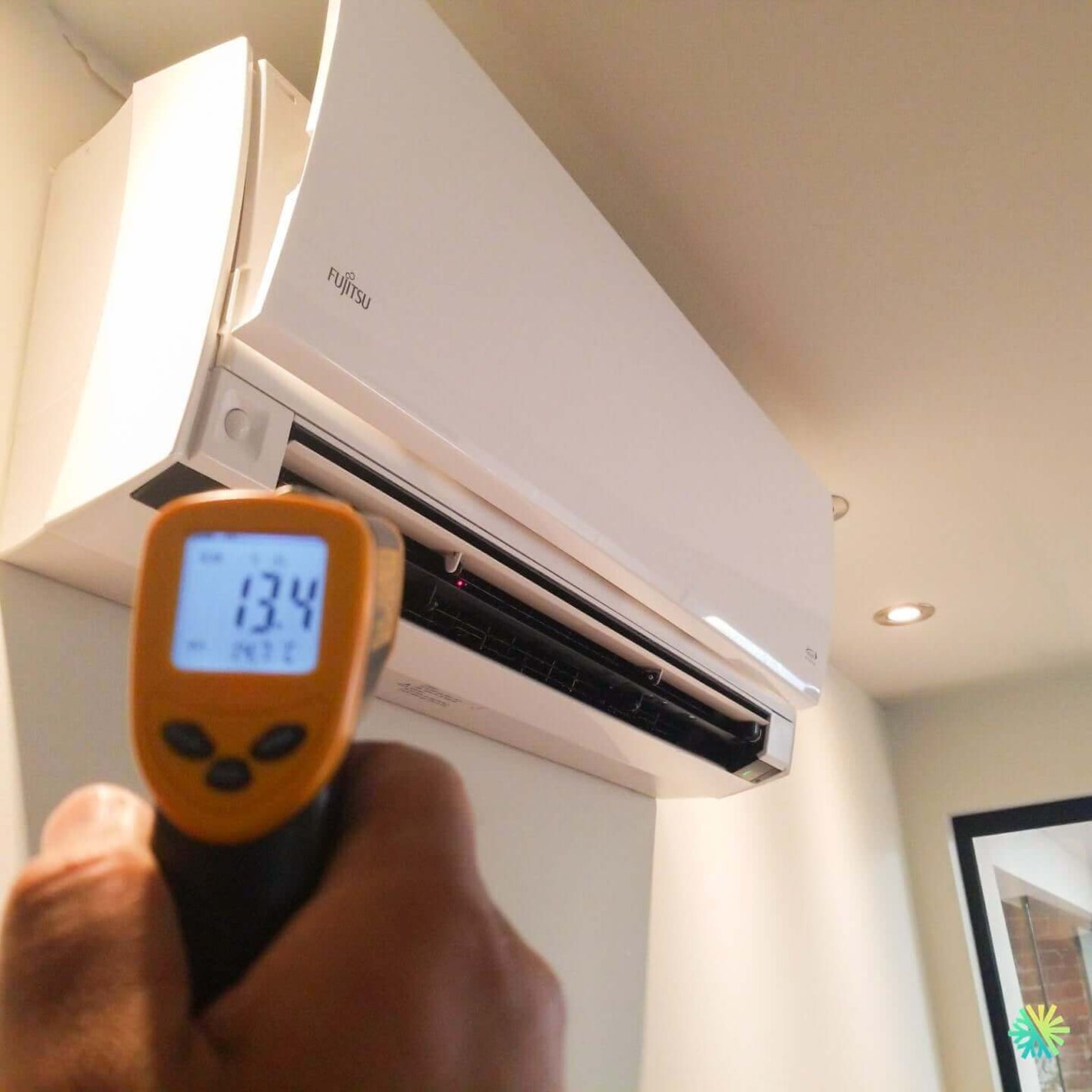Person using a digital thermometer to check temperature of a Fujitsu air conditioning unit mounted on a wall indoors.