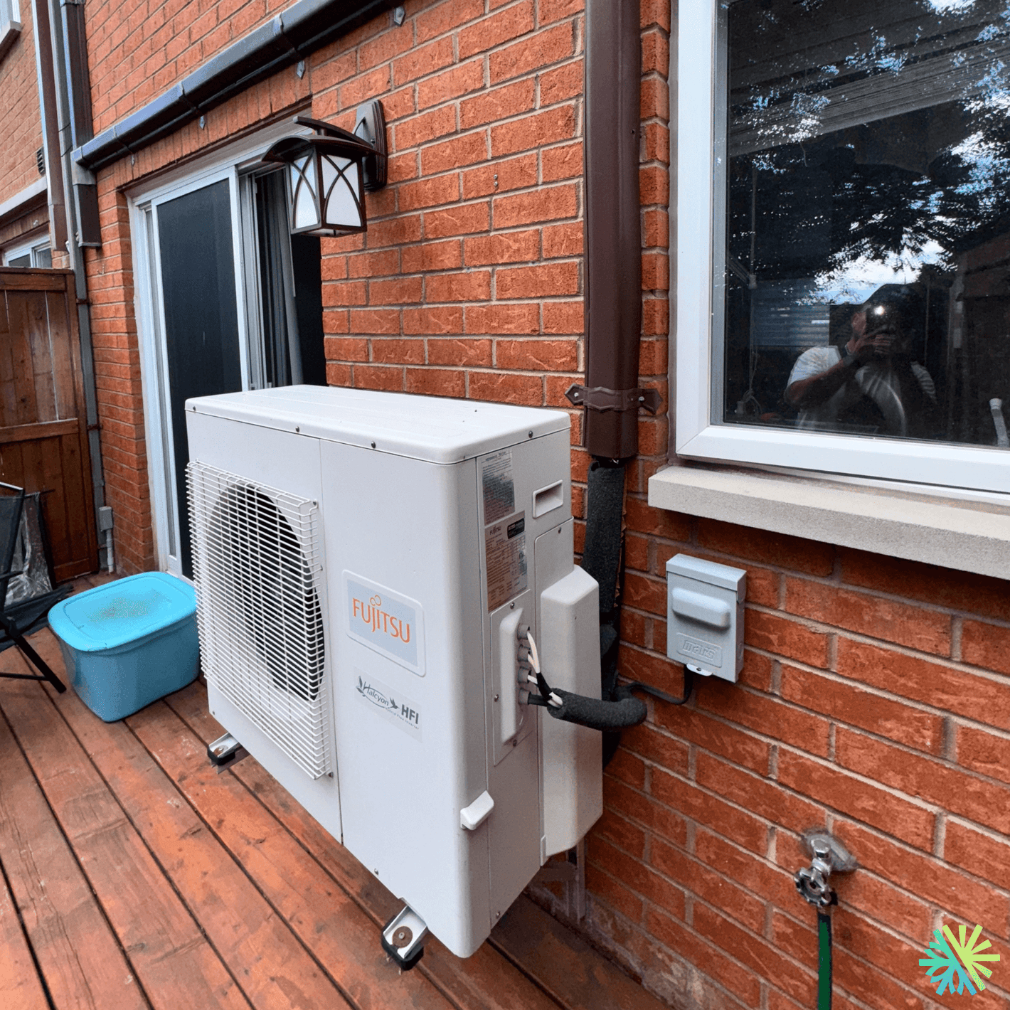 Outdoor Fujitsu air conditioning unit installed on a wooden deck outside a brick home, near a window and a water faucet.