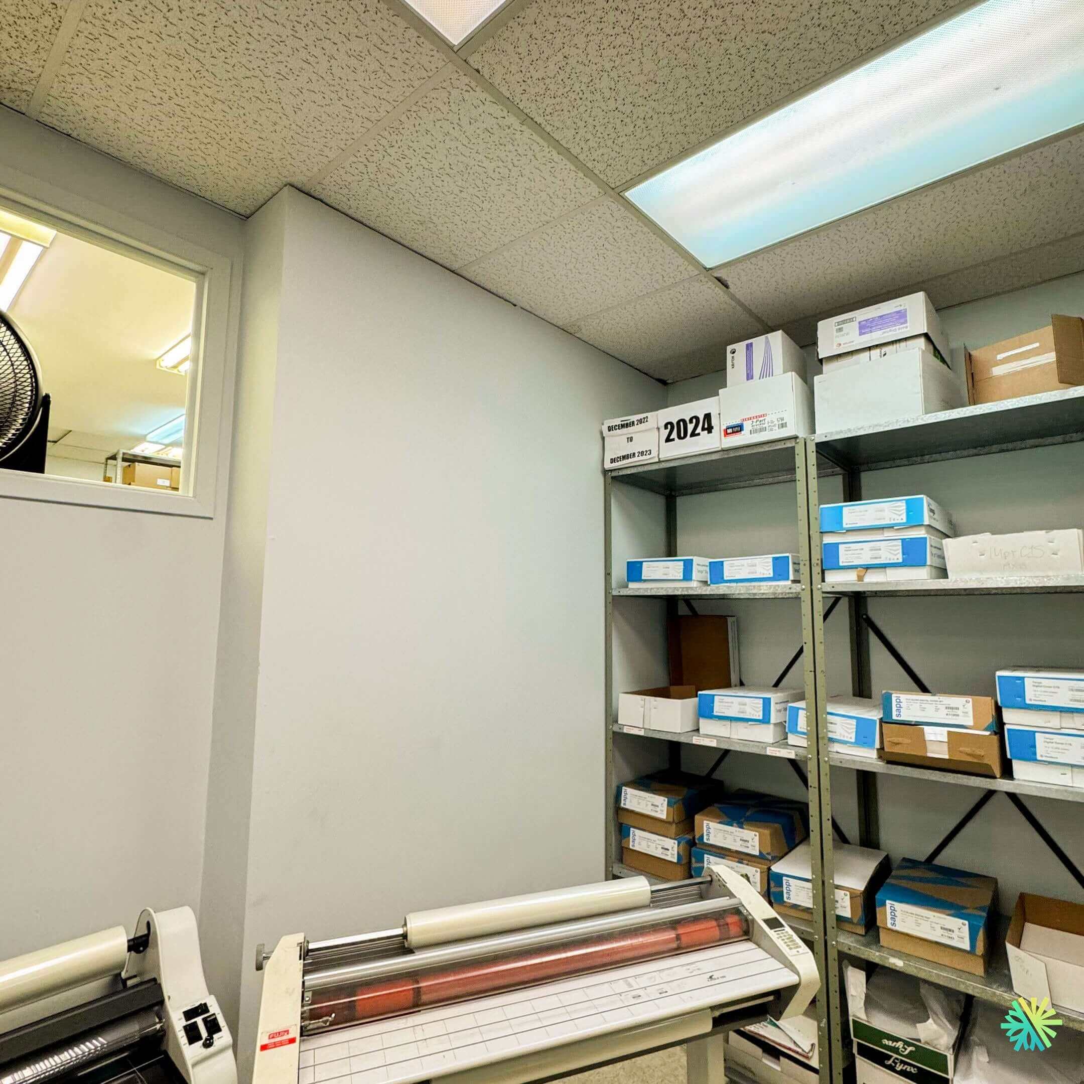Office storage room with shelves holding organized boxes and paper, featuring a laminating machine below fluorescent lighting.