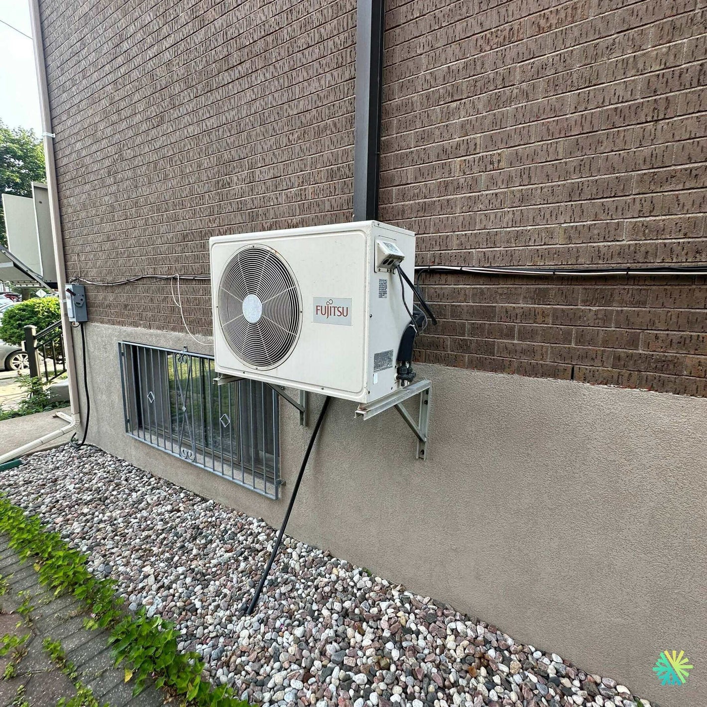 Outdoor Fujitsu air conditioning unit installed on a brick wall above a bed of decorative stones and greenery.