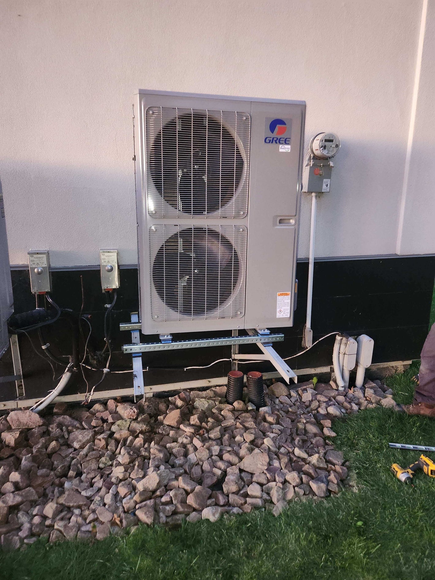 Outdoor air conditioning unit installed on a metal stand beside a building wall, surrounded by rocks and grass.