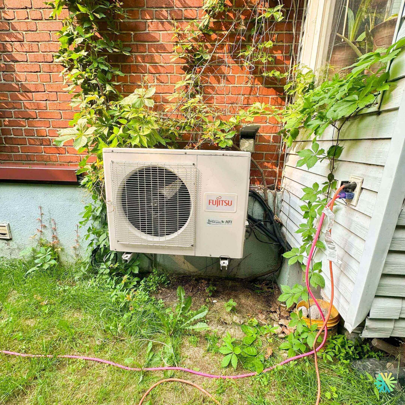 Outdoor Fujitsu air conditioning unit installed on exterior wall, surrounded by green vines and plants.