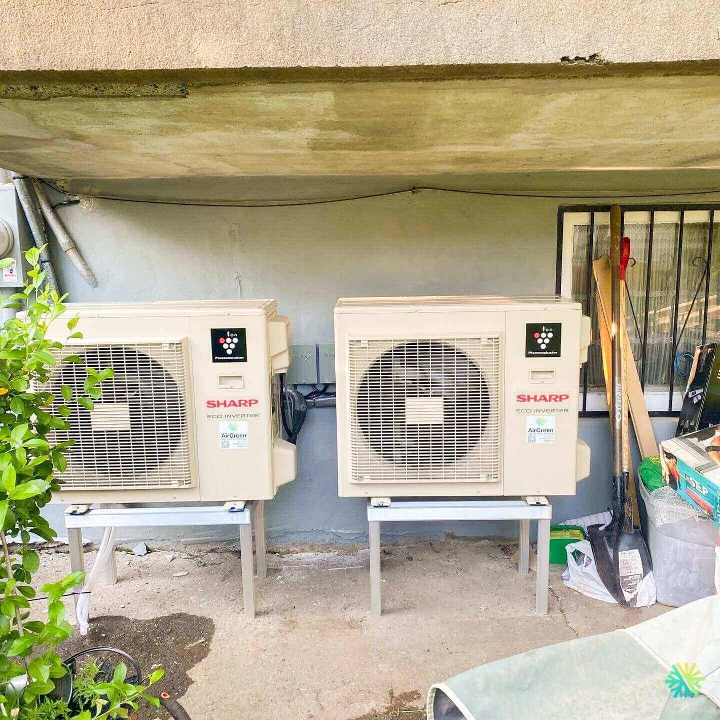 Two Sharp air conditioning units installed outside a building, surrounded by gardening tools and plants.