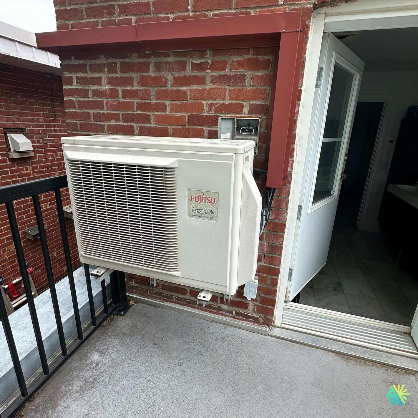 Outdoor Fujitsu air conditioning unit installed on a brick wall next to an open door on a small balcony.