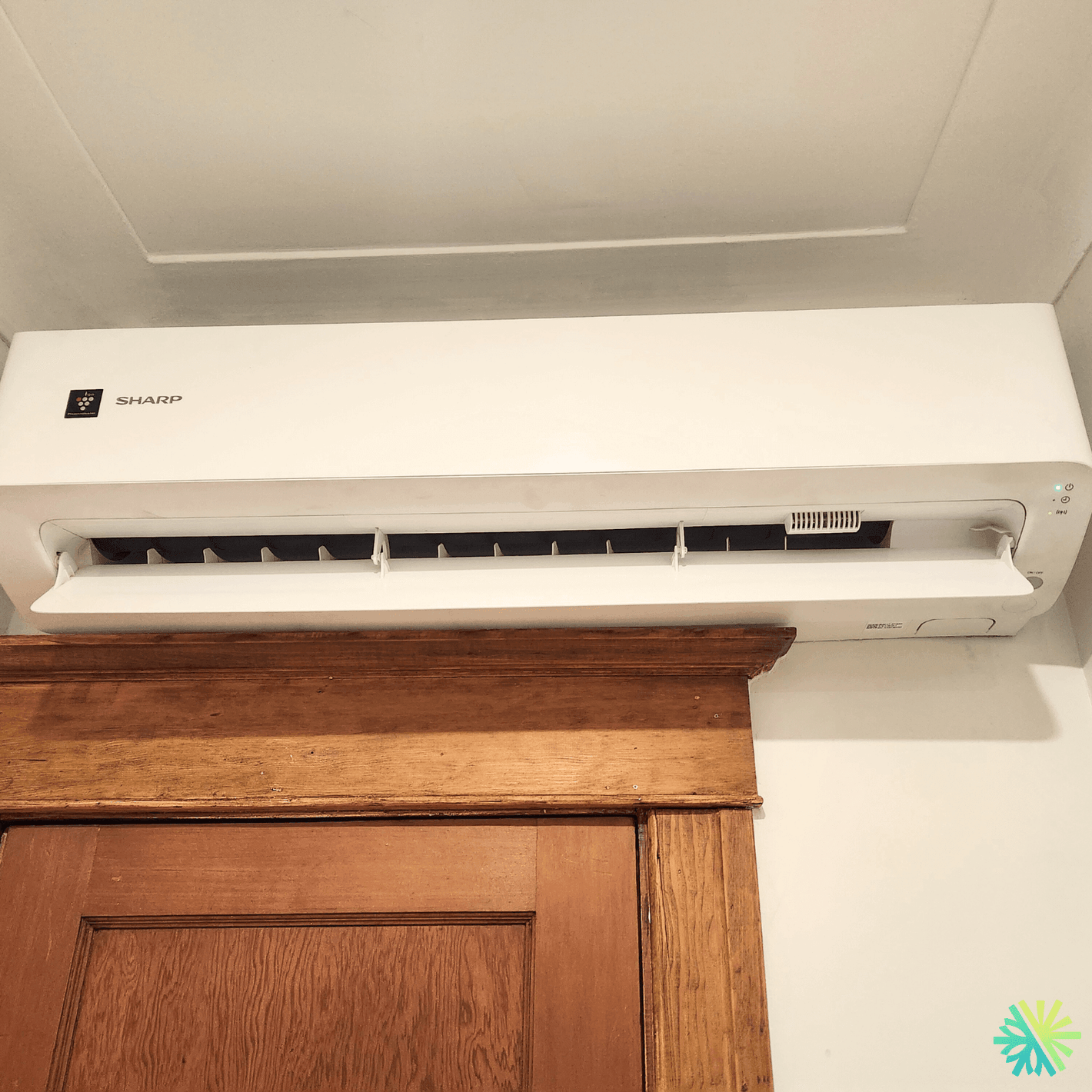 Wall-mounted Sharp air conditioner above a wooden door, cooling a room efficiently.
