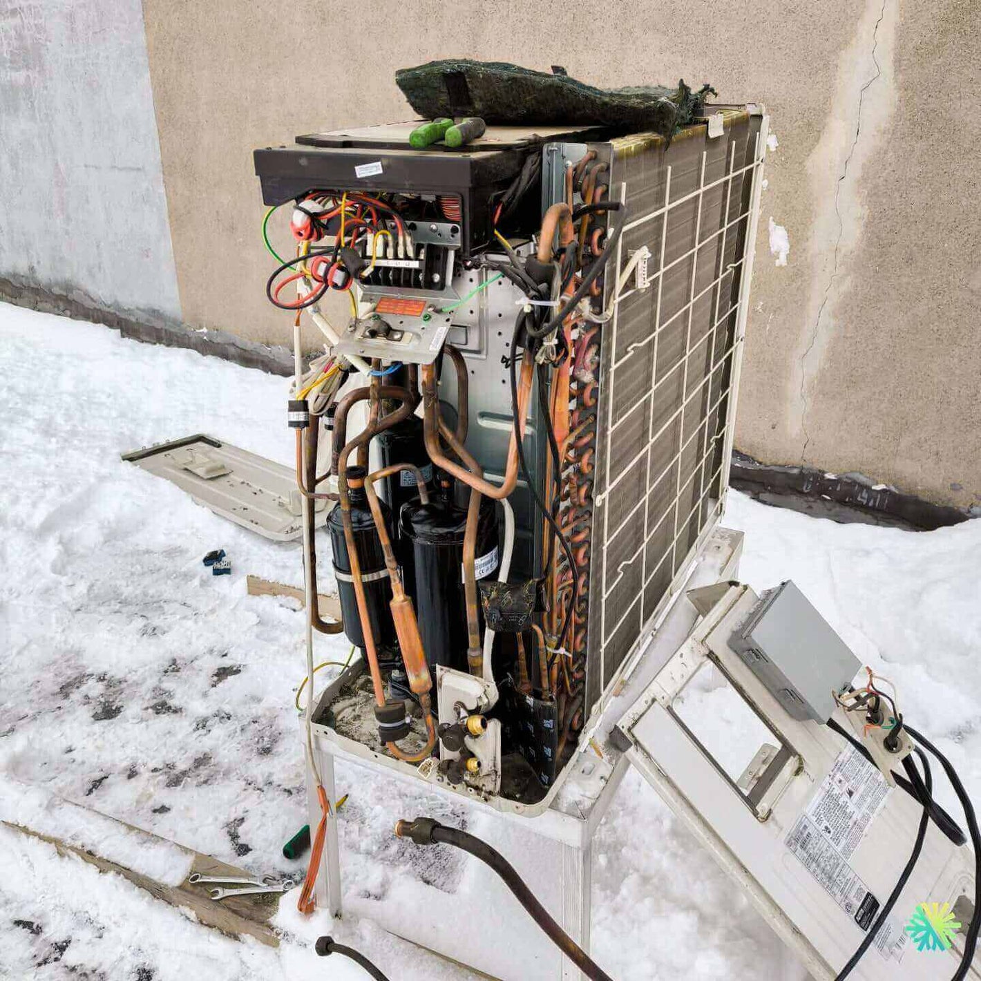 Repair of an outdoor air conditioning unit in snowy conditions, with exposed wiring and components, against a concrete wall.