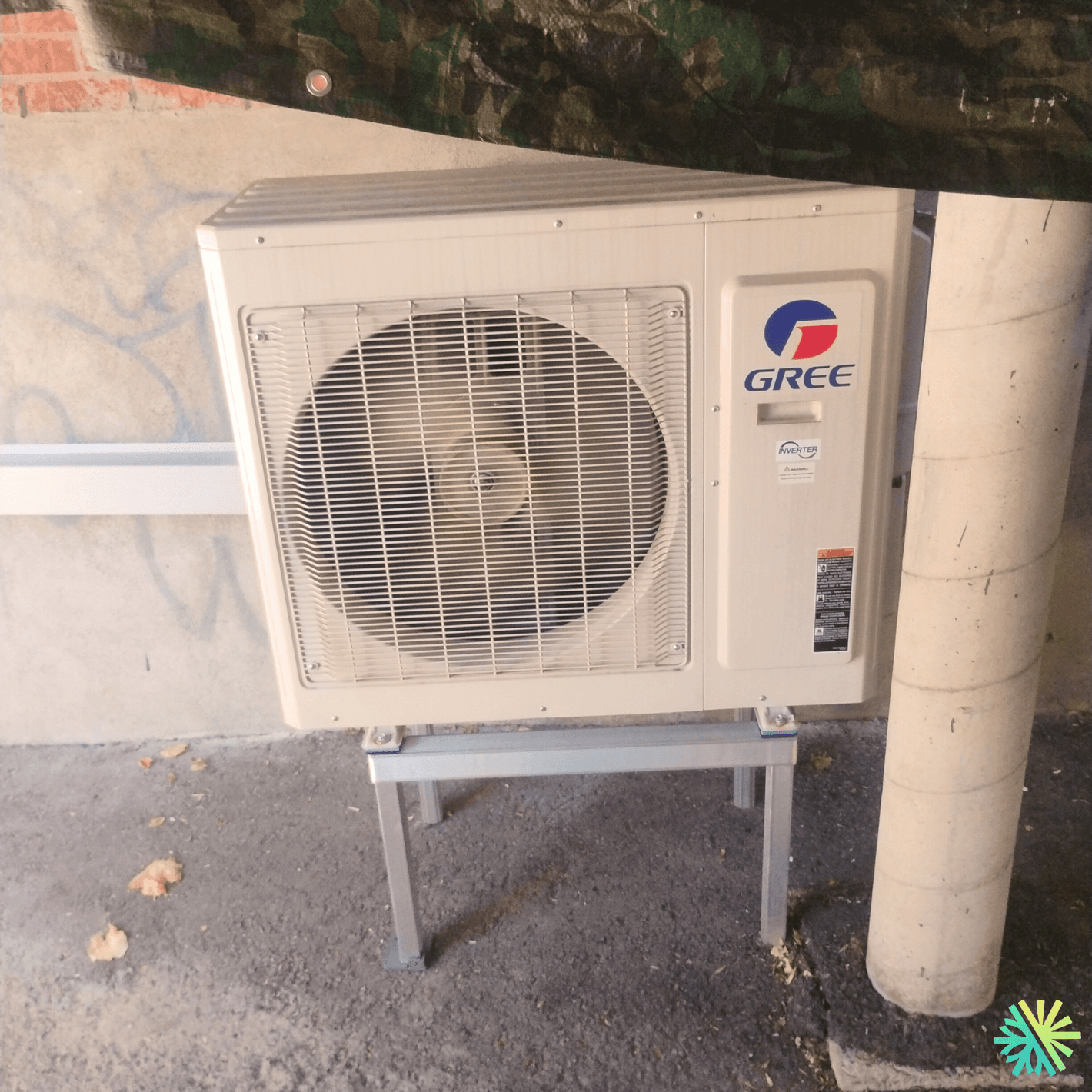 Outdoor Gree air conditioning unit mounted on a stand, positioned under a camouflage tarp next to a wall and column.