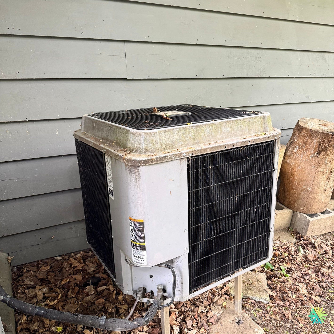 Old outdoor air conditioning unit next to a house with wooden siding, surrounded by leaves and debris.