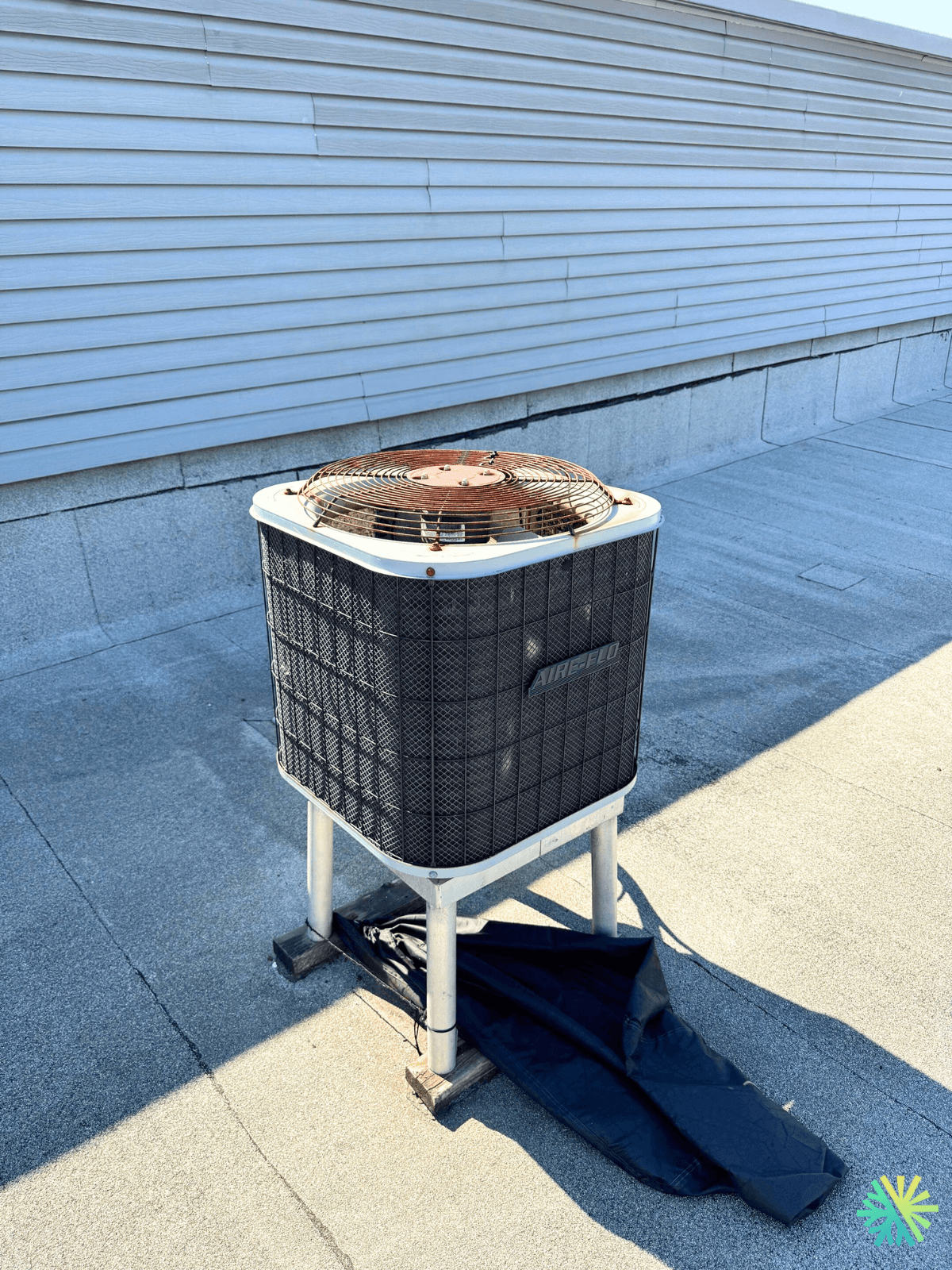 Outdoor air conditioning unit on rooftop with metal casing and fan, against a background of siding and sunlight casting shadows.
