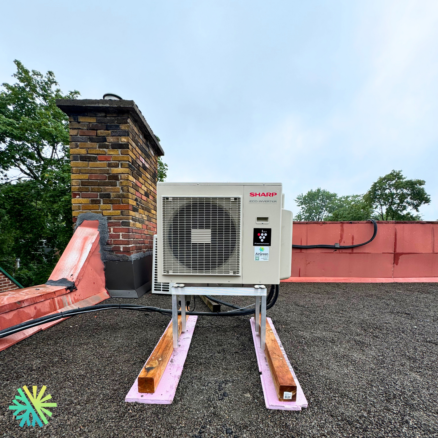 Rooftop installation of a Sharp outdoor unit with a brick chimney and lush greenery in the background.