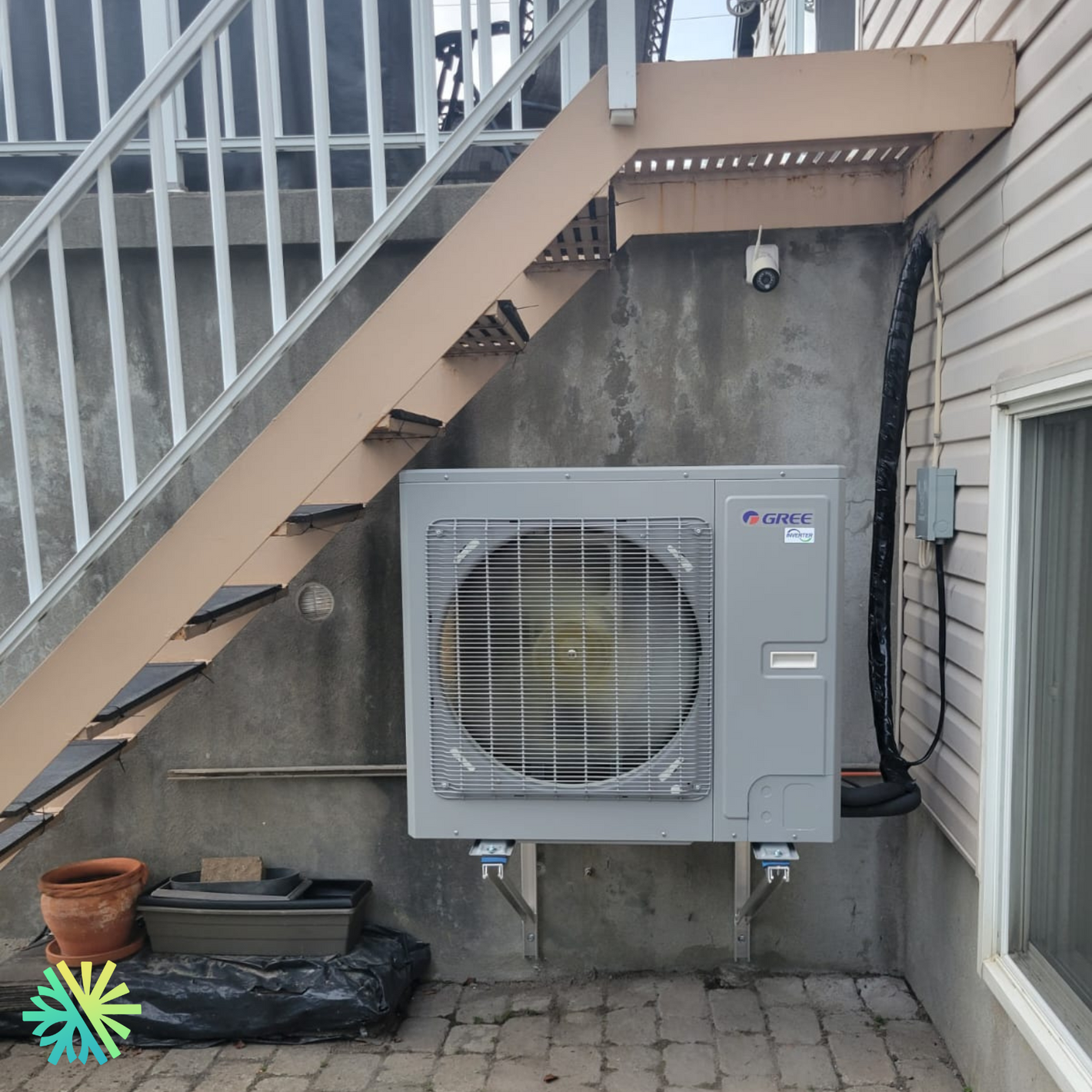 Outdoor Gree AC unit installed under stairs on a concrete wall, next to a flower pot on a paved patio.