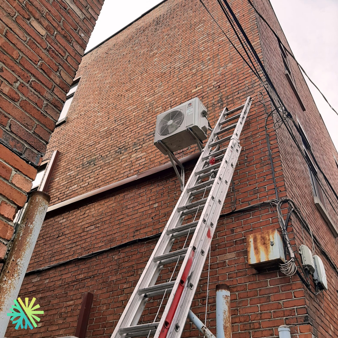 Ladder leaning against a brick building, leading to an outdoor air conditioning unit installed high on the wall.