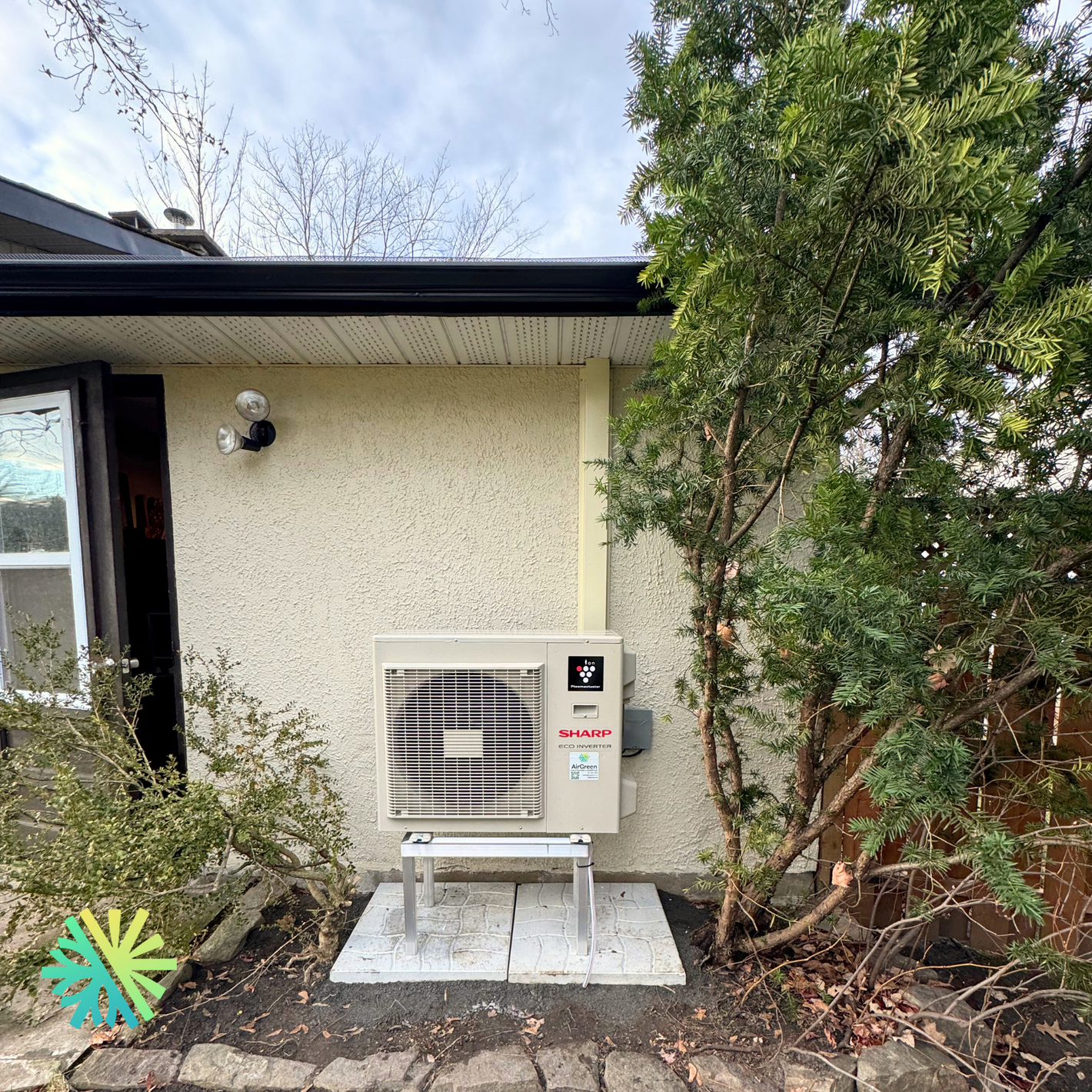 Outdoor air conditioning unit installed on a residential house wall, surrounded by trees and shrubs.