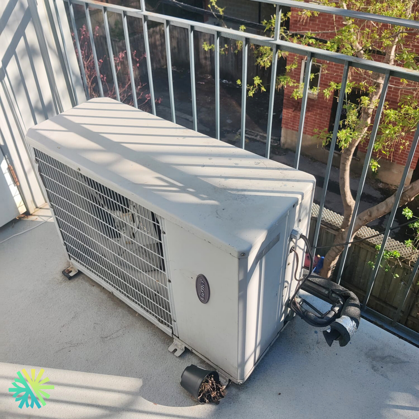 Outdoor air conditioning unit on a sunny balcony with metal railing and tree branches in the background.