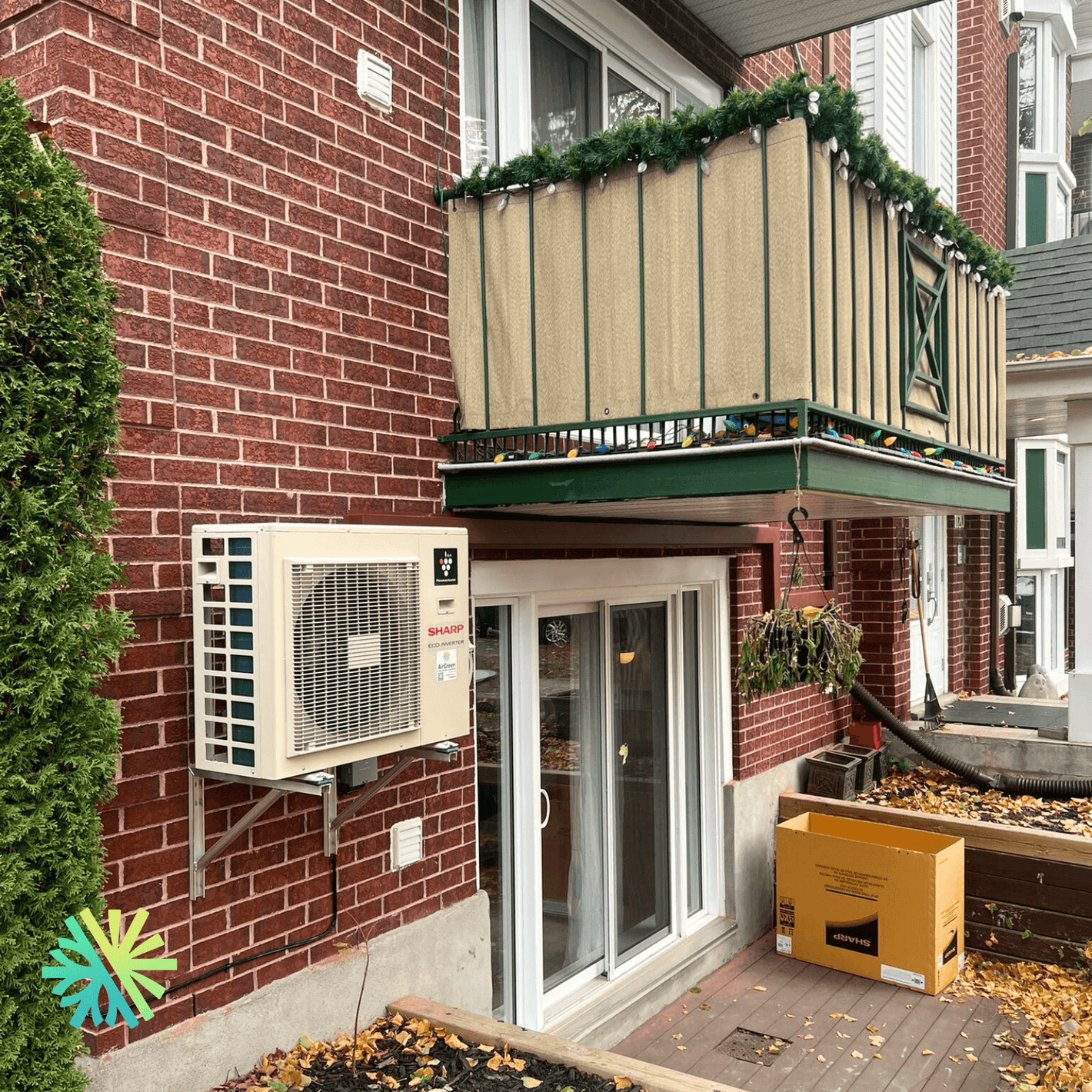Outdoor air conditioning unit installed on a brick wall of a residential building next to sliding glass doors and a balcony with decorations.